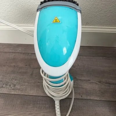 Handheld teal and white fabric steamer with brush attachment and wrapped cord.