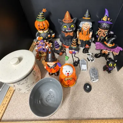 Collection of Halloween-themed decor items including painted ceramic figures of witches, black cats, and jack-o'-lanterns alongside...