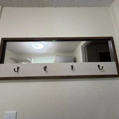 Modern wooden wall-mounted coat rack featuring an integrated upper mirror, light fabric backing, and four metal hooks.