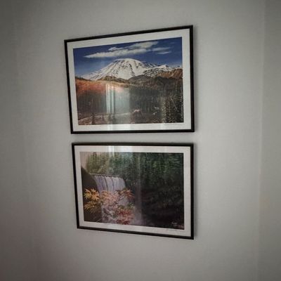 Pair of framed nature landscape prints depicting a snow-capped mountain and a forest waterfall, both mounted in matching black frames...