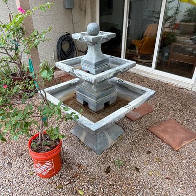 Three-tiered square concrete water fountain with a finial ball, set in a gravel patio area.