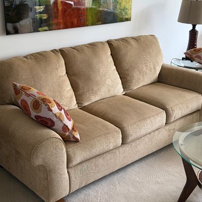 Contemporary beige upholstered three-cushion sofa featuring rolled arms and wooden feet, styled with a patterned throw pillow.