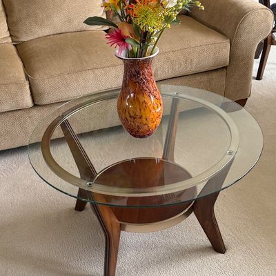 Round modern glass-top accent table with wooden legs and metal trim, holding a multicolored art glass vase filled with artificial flowers.