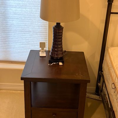 Dark wood nightstand featuring a single lower drawer, open shelf storage, and surface wear, topped with a textured ceramic table lamp.