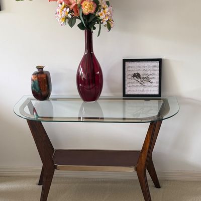 Modern glass-top console table with wooden legs and lower shelf, styled with a tall maroon vase of faux flowers, a ceramic vessel, and...