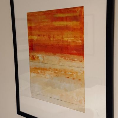 Framed contemporary abstract print featuring warm orange, yellow, and beige color fields, presented in a black frame with wide white...