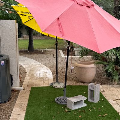 Two outdoor patio umbrellas, one pink and one yellow, set in weighted bases outdoors on an artificial turf pad.