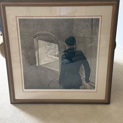 Large framed contemporary art print depicting a figure in a coat and beanie looking out a window, presented in a wooden frame with...