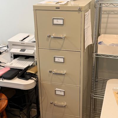Beige four-drawer OfficeMax metal vertical filing cabinet with silver handles and label holders, flanked by a printer and wire shelving...