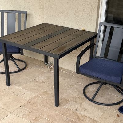 Modern outdoor patio set featuring a square wood-look slatted table with an umbrella hole and two black metal swivel armchairs with blue...