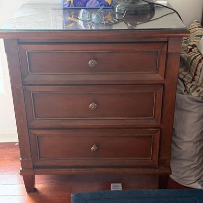 Traditional wooden three-drawer nightstand featuring paneled drawer fronts, antique brass knobs, and a protective glass glass top.