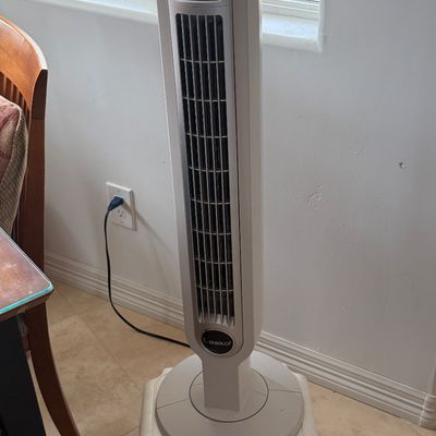 Lasko modern oscillating tower fan in silver and white, shown powered on and sitting atop a plastic stool.