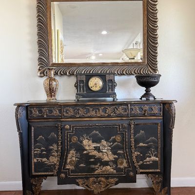 Ornate black and gold French Chinoiserie chest of drawers with a large gilded spiral-frame wall mirror, a mantle clock, and decorative...