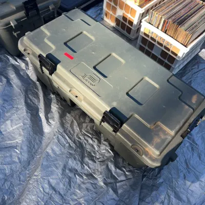 Sale Photo Thumbnail #3: Large olive drab Plano molded plastic storage trunk with heavy-duty latches, shown alongside other outdoor equipment and vinyl record crates on a tarp.