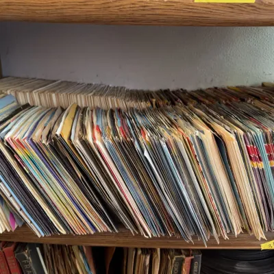 Sale Photo Thumbnail #103: Large collection of vintage 45 RPM vinyl records in paper sleeves packed onto a wooden shelf.