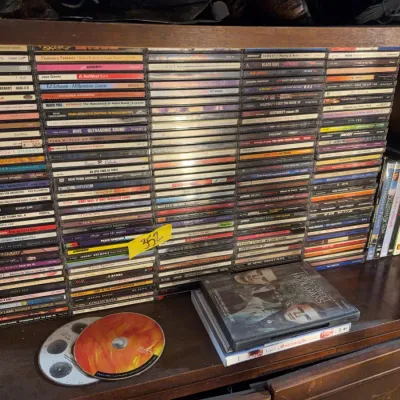 Sale Photo Thumbnail #160: Large collection of music CDs in jewel cases stacked on a wooden shelf, alongside various DVD movies.