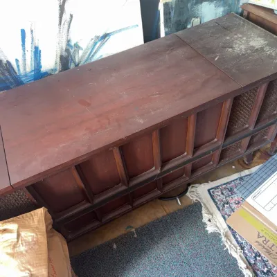 Sale Photo Thumbnail #178: Vintage mid-century modern wooden stereo console with integrated speaker grilles, showing wear and surface dust.