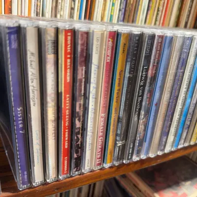 Sale Photo Thumbnail #138: Large collection of compact discs in jewel cases lined up on a wooden shelf, featuring various pop, rock, and classical albums.