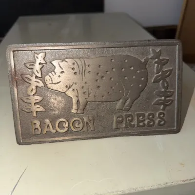 Sale Photo Thumbnail #189: Cast iron bacon press featuring a raised spotted pig motif and leaf accents, with 'BACON PRESS' lettering across the bottom.