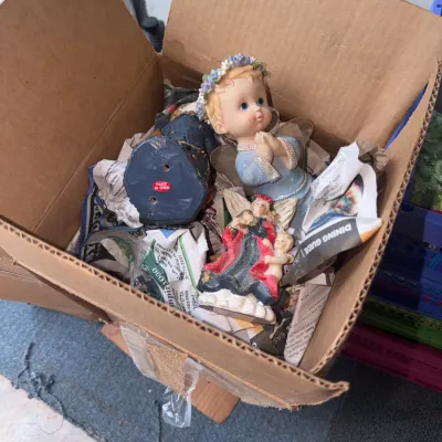 Sale Photo Thumbnail #172: Cardboard box containing a assortment of decorative angel figurines packed in newspaper.
