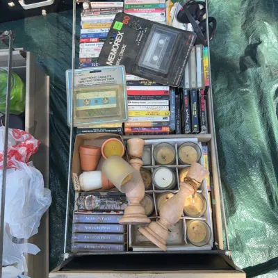 Sale Photo Thumbnail #48: Open metal drawer filled with vintage cassette tapes, a Memorex blank cassette in original packaging, a tape head demagnetizer, small candles, and wooden candleholders.