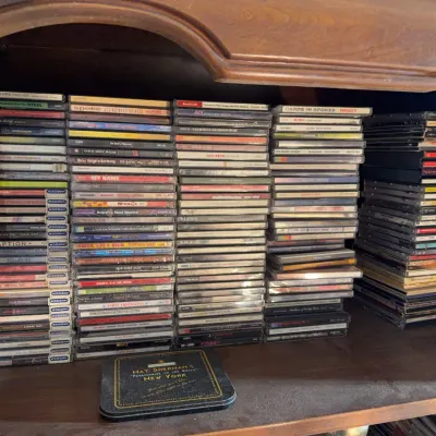 Sale Photo Thumbnail #161: Large collection of music CDs in jewel cases, neatly organized in stacks inside a wooden entertainment center, alongside a vintage Nat Sherman tin.