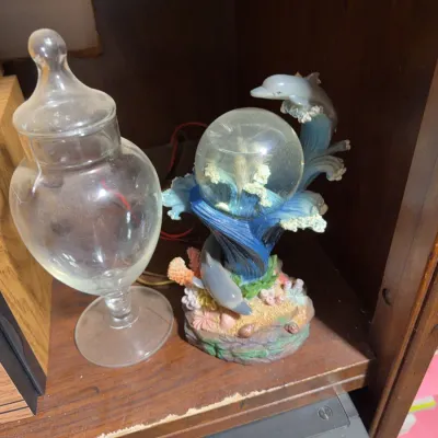 Sale Photo Thumbnail #169: Clear glass apothecary jar with finial lid sitting next to a decorative resin dolphin water globe figurine on a wooden shelf.