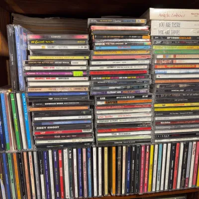 Sale Photo Thumbnail #125: Large collection of assorted music CDs stacked on wooden shelving, featuring various genres and artists from the 1990s and 2000s.