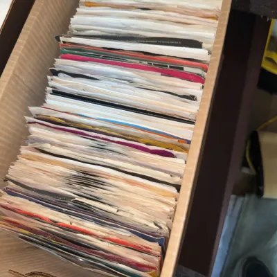 Sale Photo Thumbnail #181: Cardboard storage box filled with a collection of vintage 45 RPM vinyl records in paper sleeves.