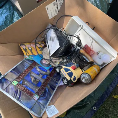 Sale Photo Thumbnail #50: Cardboard box containing a mixture of household items, including a leaded stained glass panel, toy police car, electronic cords, and aerosol cans.