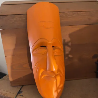 Sale Photo Thumbnail #156: Hand-carved wooden decorative face mask painted bright orange, resting against a wooden trunk.