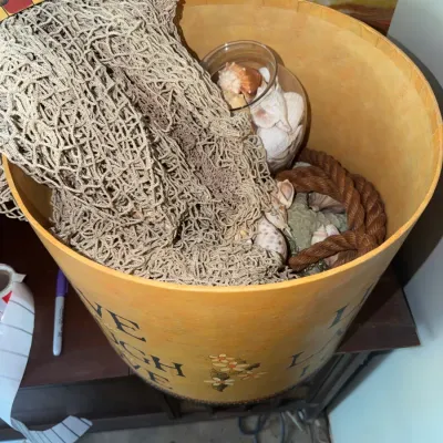Sale Photo Thumbnail #188: Large painted decorative round storage container filled with nautical decor, including fishnet, sea shells, a glass jar, and rope.