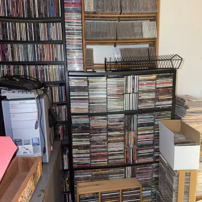 Sale Photo Thumbnail #191: Large media collection featuring hundreds of compact discs in various wooden and wire storage racks alongside a vintage computer tower.