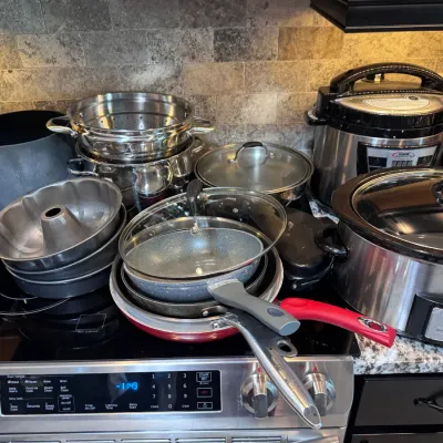 Sale Photo Thumbnail #129: Collection of modern kitchen cookware and small appliances, including a Crock-Pot slow cooker, pressure cooker, stacked frying pans with glass lids, bundt pans, and stainless steel steamers.