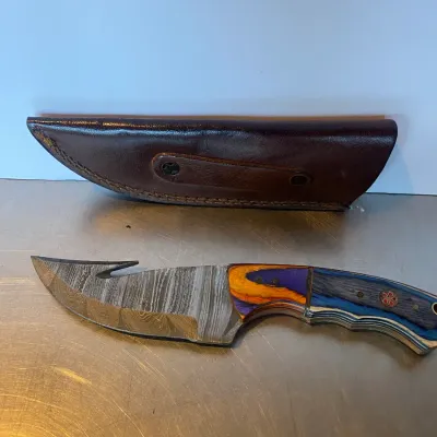 Sale Photo Thumbnail #58: Custom Damascus steel skinning knife with a gut hook, colorful composite handle with mosaic pins, and an accompanying fitted brown leather sheath.