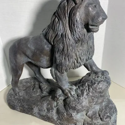Sale Photo Thumbnail #116: Cast metal or resin statue of a standing lion with a heavy mane, resting one paw on a rocky base, featuring a dark patinated finish.