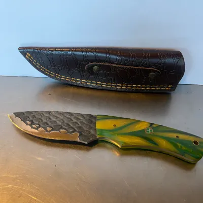 Sale Photo Thumbnail #64: Custom fixed-blade hunting knife featuring a textured scale-pattern steel blade and a vibrant yellow and green swirl resin handle, paired with a fitted dark brown leather sheath.