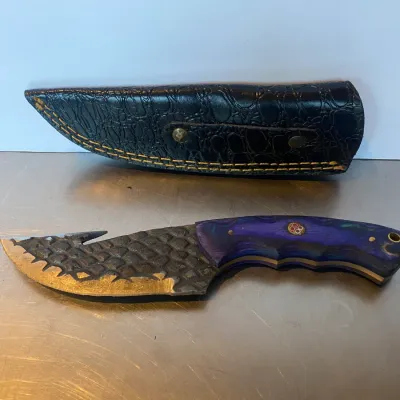Sale Photo Thumbnail #65: Custom fixed-blade hunting knife featuring a textured dragon-scale steel finish, gut hook, and purple resin handle, paired with a black faux-croc leather sheath.