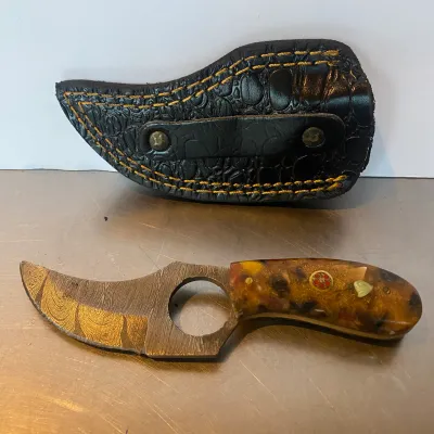 Sale Photo Thumbnail #100: Custom Damascus steel skinning knife with a finger hole, decorative composite resin handle, and matching embossed black leather sheath.