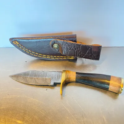 Sale Photo Thumbnail #82: Fixed-blade Damascus steel knife with a brass guard and contoured horn and wood handle, shown with an alligator-embossed leather sheath.