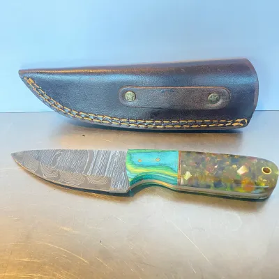 Sale Photo Thumbnail #79: Fixed blade Damascus steel knife with a colorful resin and laminate handle, accompanied by a stitched brown leather sheath.