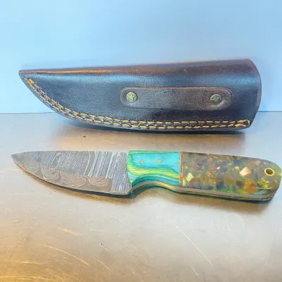 Sale Photo Thumbnail #52: Fixed-blade Damascus steel knife featuring a vibrant composite resin and laminated wood handle, presented alongside its fitted brown leather sheath.
