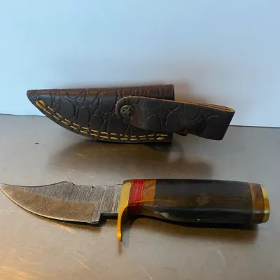 Sale Photo Thumbnail #66: Handcrafted Damascus steel fixed-blade hunting knife featuring a brass guard, wood and horn segmented handle, and an embossed brown leather sheath.