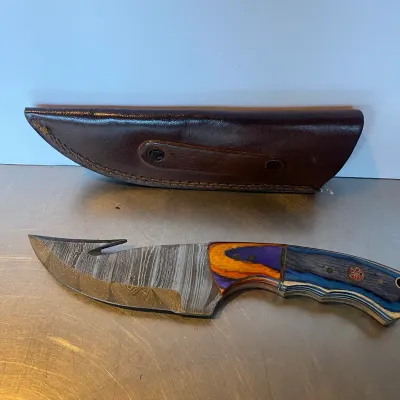 Sale Photo Thumbnail #85: Custom Damascus steel skinning knife with gut hook, featuring a colorful wood and composite handle, complete with a fitted brown leather sheath.