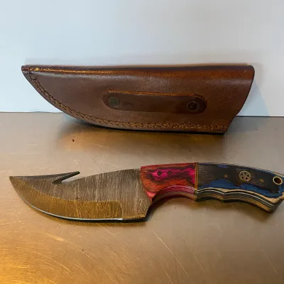 Sale Photo Thumbnail #99: Custom Damascus steel hunting knife featuring a gut hook and a multi-colored wood laminate handle, accompanied by a fitted brown leather sheath.