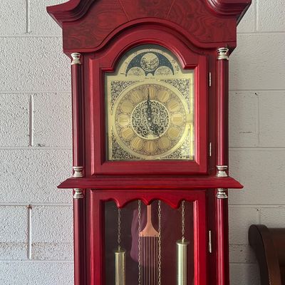 Sale Photo Thumbnail #3: Traditional grandfather clock in a painted red wood casing, featuring an ornate brass face with Roman numerals and visible pendulum and weights.