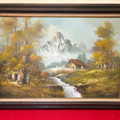 Framed oil painting depicting a rustic cabin landscape with snow-capped mountains and a stream, signed in the lower right corner.