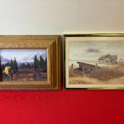 Pair of framed Western-themed art prints, including a mountain landscape in a thick oak frame and a rural homestead scene in a brass-tone...