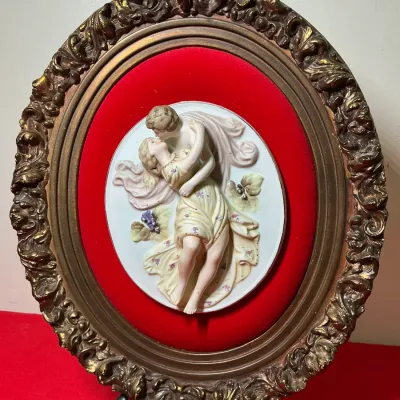 Vintage oval framed 3D bisque porcelain plaque featuring a romantic classical scene, mounted on red velvet inside an ornate gilded frame.