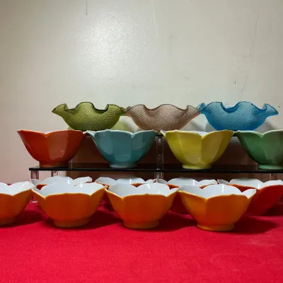 Collection of colorful vintage art glass and porcelain flower-form dessert bowls, displayed on tiered riser shelves.